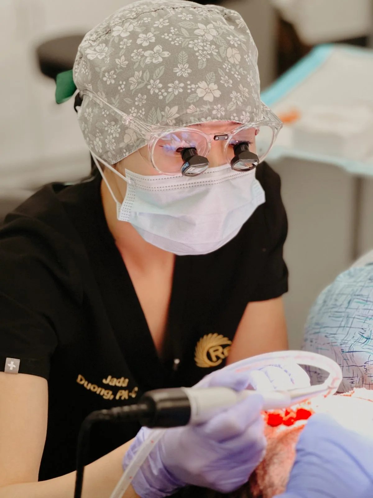 Dermatologist wearing magnifying loupes and protective gear examining a patient's skin lesion with a handheld device